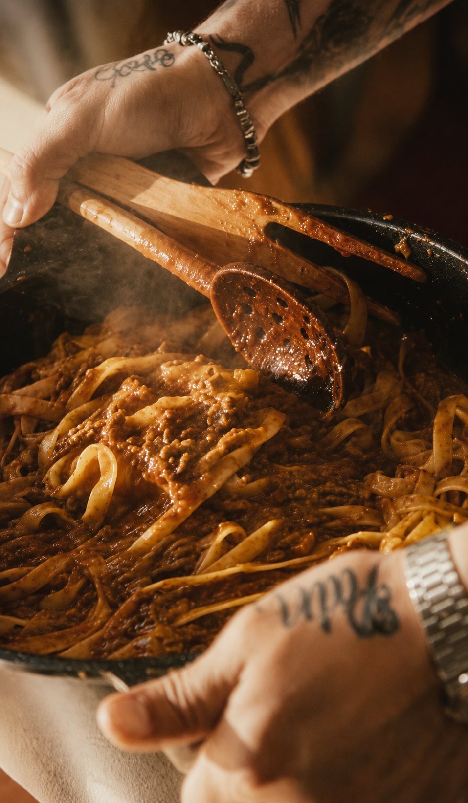 Hands lifting steaming fresh tagliatelle and ragù from a black cast-iron pan with a wooden spoon